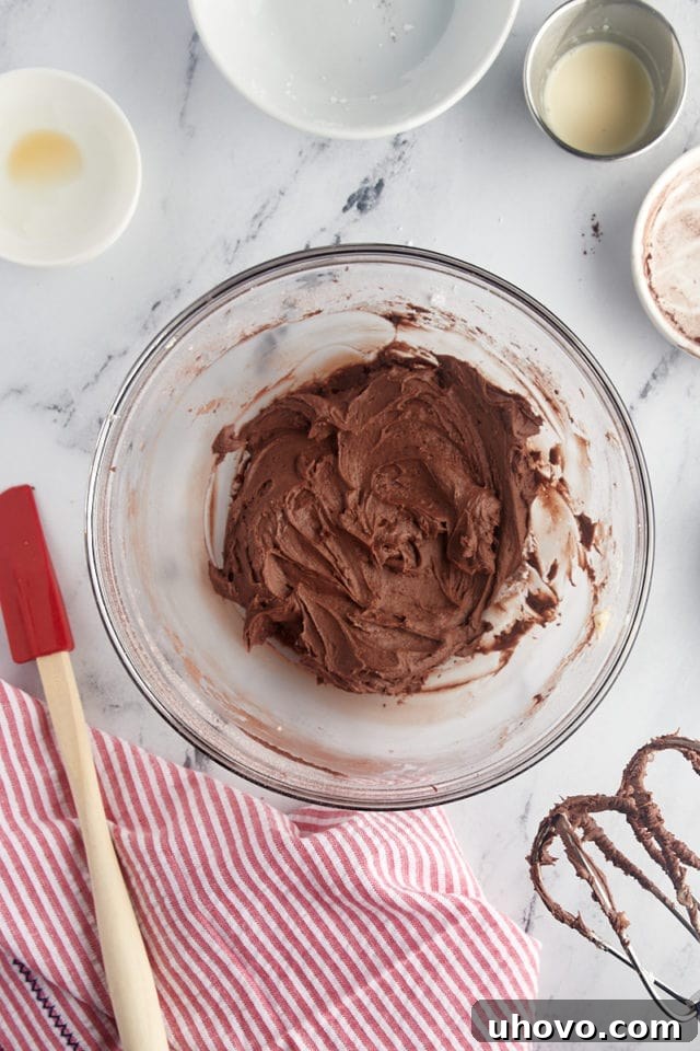 A close-up, overhead view of velvety smooth chocolate buttercream frosting, perfectly whipped and awaiting its moment to adorn the cookie cake, resting in a glass mixing bowl.