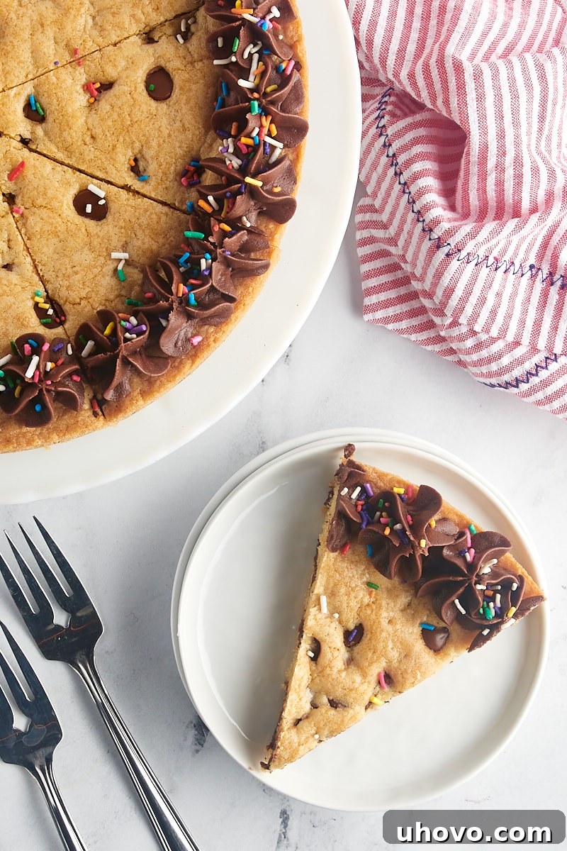 An inviting overhead view of a single slice of chocolate chip cookie cake presented on a white plate, perfectly complemented by the remaining cake resting on a white cake stand nearby.