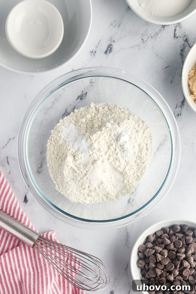 An overhead view of a clear glass mixing bowl filled with flour, cornstarch, baking soda, and salt, perfectly portioned and ready to be whisked together.