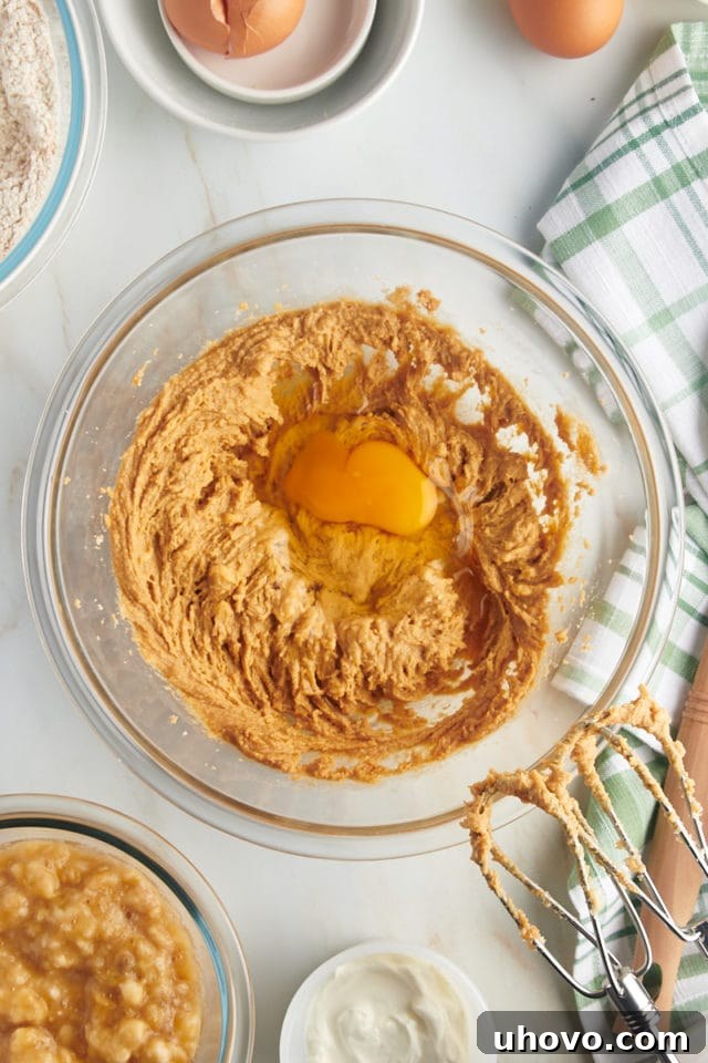 Overhead view of an egg freshly cracked into the mixing bowl, joining the creamed butter, sugar, and peanut butter, ready to be incorporated.