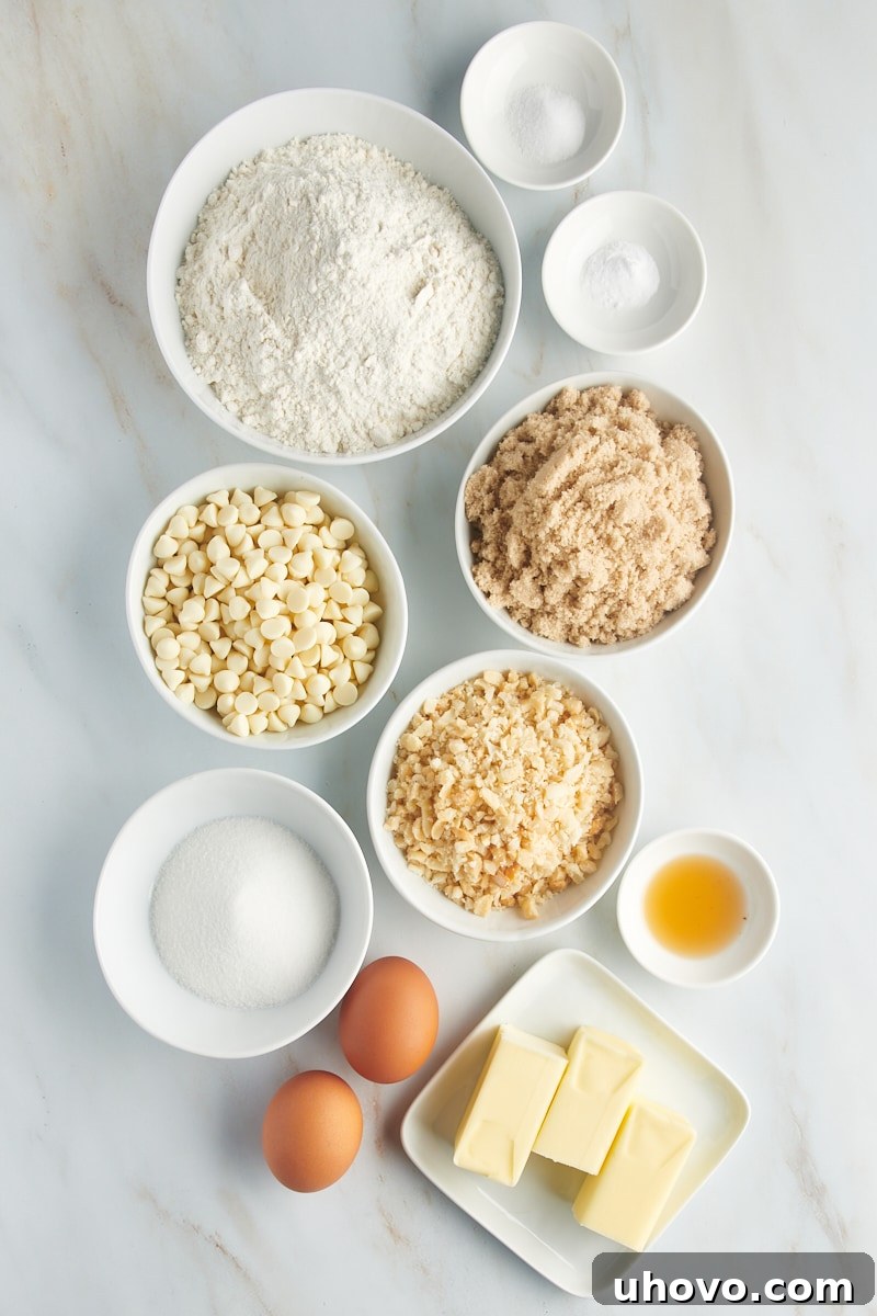 Overhead view of individual ingredients for white chocolate macadamia nut cookies laid out on a light surface, including flour, sugars, butter, eggs, chocolate chips, and macadamia nuts.