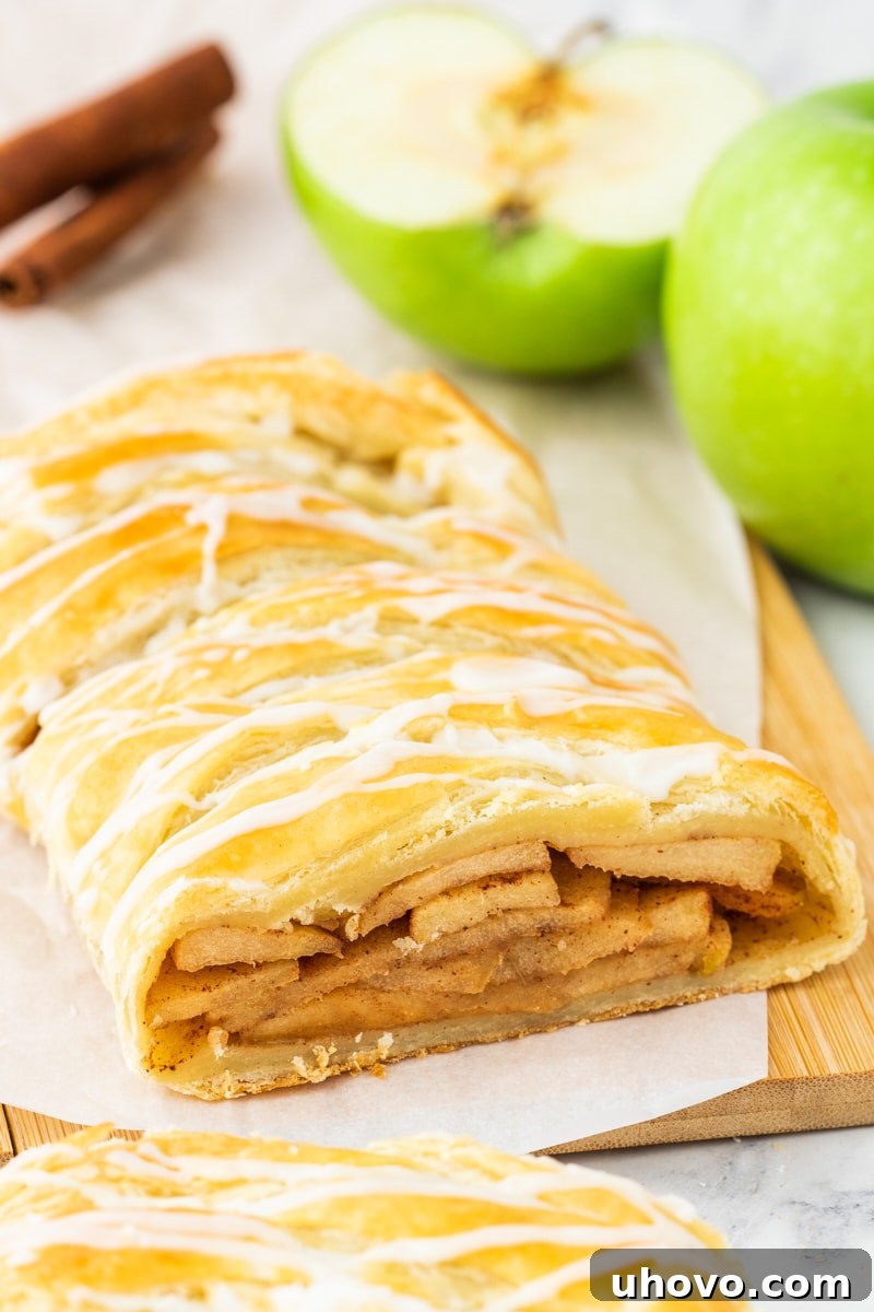 Delicious homemade apple strudel on parchment paper on a wooden cutting board, with a sliced green apple and cinnamon sticks in the background, highlighting the fall flavors.