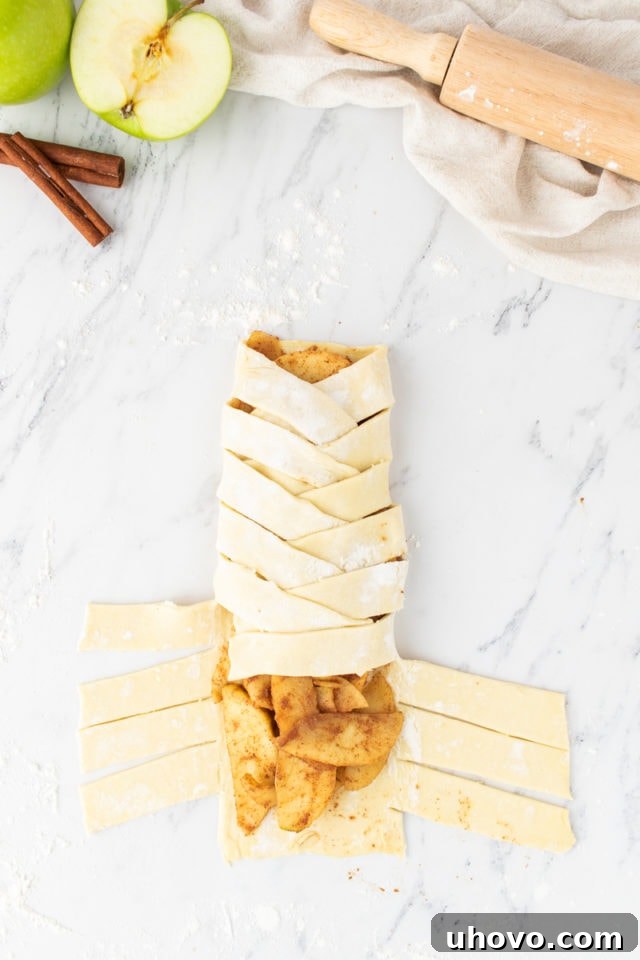 Overhead view of a partially assembled apple strudel, with pastry strips being folded over the apple filling in an alternating pattern.