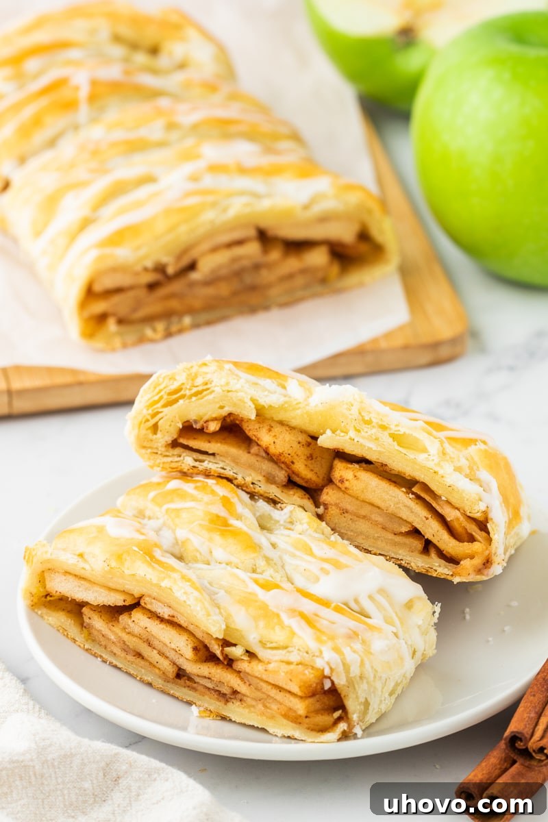 Two alluring slices of apple strudel on a white plate, with more strudel and fresh apples subtly blurred in the background, creating a cozy autumn scene.