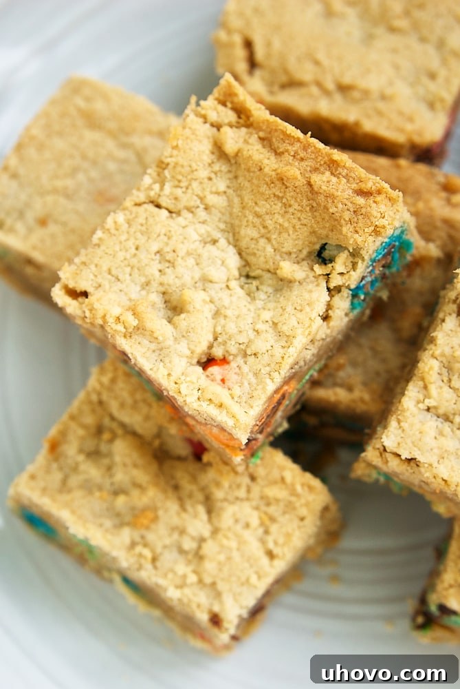 Overhead view of neatly stacked Peanut Butter Cookie Bars on a white surface, highlighting their golden-brown edges and abundance of colorful M&Ms throughout each bar.