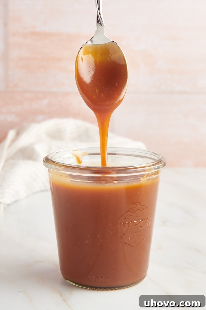 A glass jar of glistening homemade caramel sauce with a spoon resting over it, caramel dripping tantalizingly.