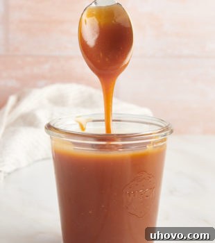 A glass jar of caramel sauce with a spoon over it with caramel dripping off of it