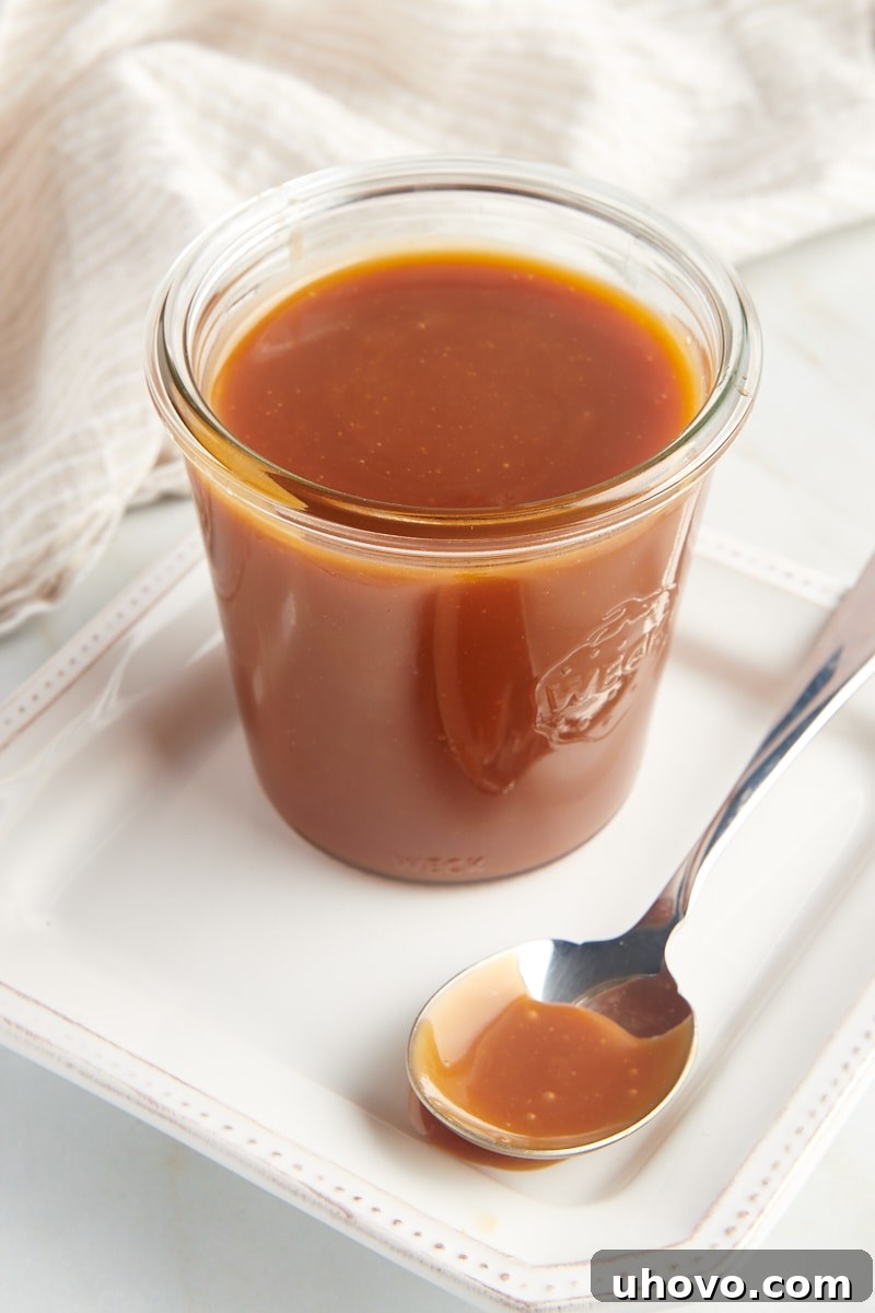 A charming glass jar of homemade caramel sauce sits on a pristine white plate, accompanied by a spoon, ready to be enjoyed.