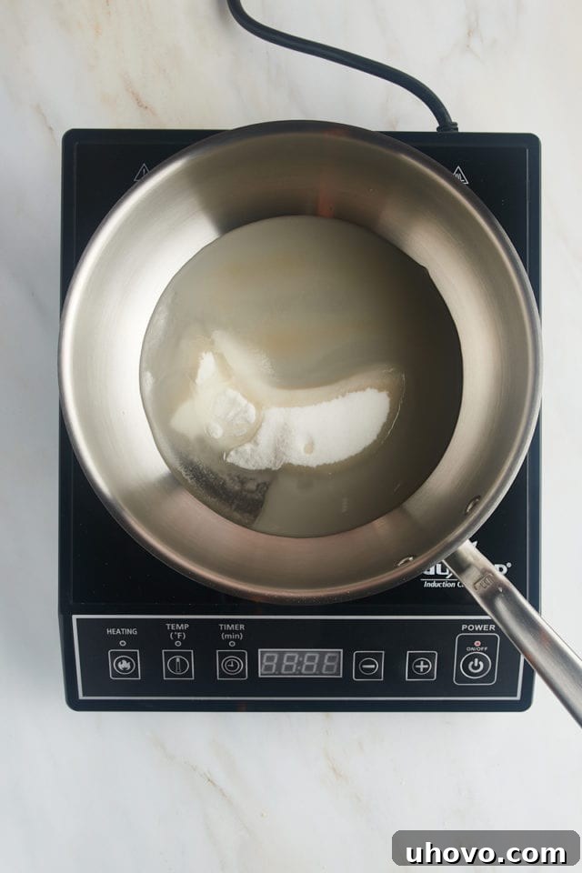 Overhead view of granulated sugar, water, and corn syrup being combined in a saucepan on an induction cooktop.