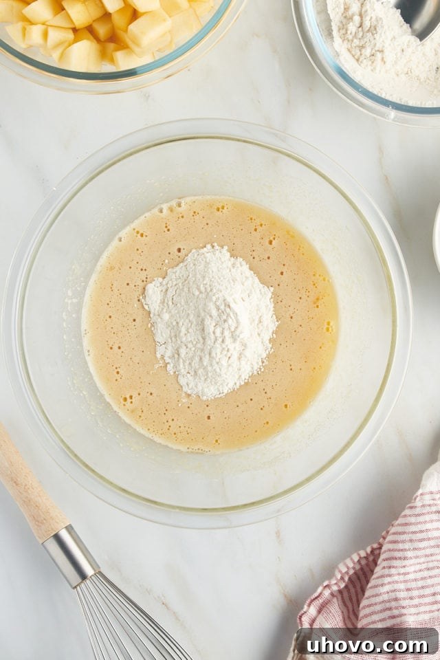 An overhead view showing a portion of the dry ingredients being carefully added to the wet cake batter in a glass mixing bowl, initiating the next stage of blending for French apple cake.
