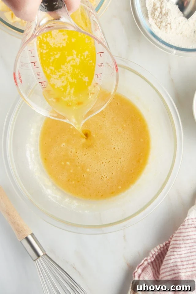 A close-up, overhead shot of warm, melted butter being poured into the partially mixed apple cake batter in a glass bowl, contributing richness and moisture to the French apple cake.