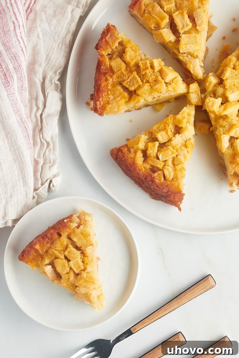 An inviting overhead view showcasing a single slice of rustic French apple cake presented on a pristine white plate, with the remaining golden-brown cake resting gracefully on a matching white cake stand, highlighting its simple beauty.