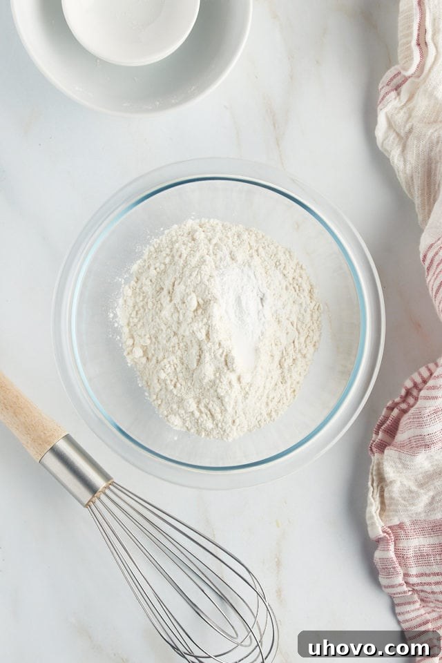 An overhead view of a clear glass mixing bowl containing precise measures of all-purpose flour, baking powder, and a pinch of salt, ready for combining to make French apple cake.