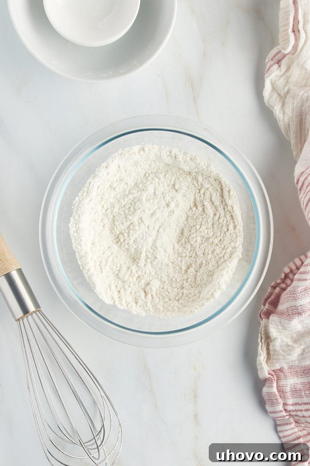 A close-up, overhead perspective of the mixed dry ingredients for French apple cake—flour, baking powder, and salt—whisked together smoothly in a glass mixing bowl.