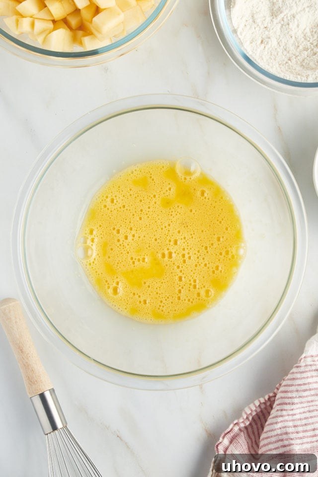 An overhead shot captures two lightly beaten eggs in a clear glass bowl, showcasing their frothy texture, ready for the next step in making French apple cake.