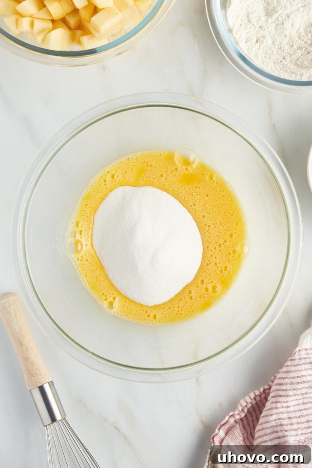 An overhead perspective shows granulated sugar being carefully added to lightly beaten eggs in a clear glass bowl, a crucial step for the French apple cake batter.