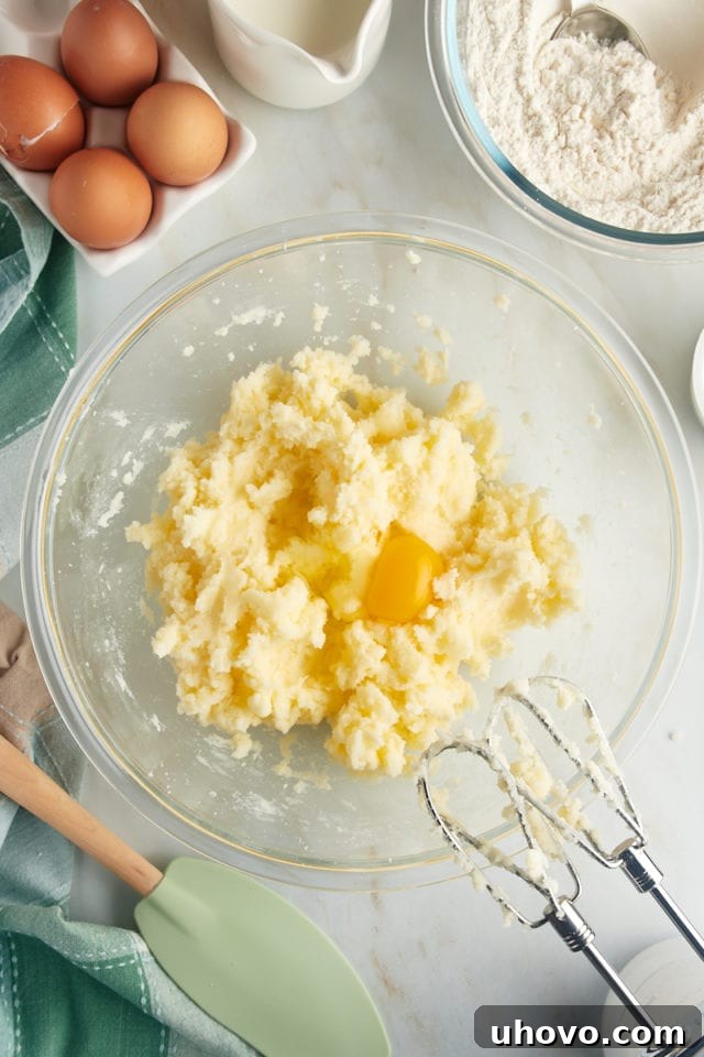 An overhead view depicting an egg being carefully added to the creamed mixture of butter and sugar in a glass mixing bowl, an essential step in building the cake batter's structure.