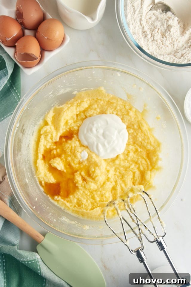 An overhead shot revealing the addition of sour cream, vanilla extract, and almond extract into the partially mixed cake batter within a glass mixing bowl, infusing it with moisture and flavor.