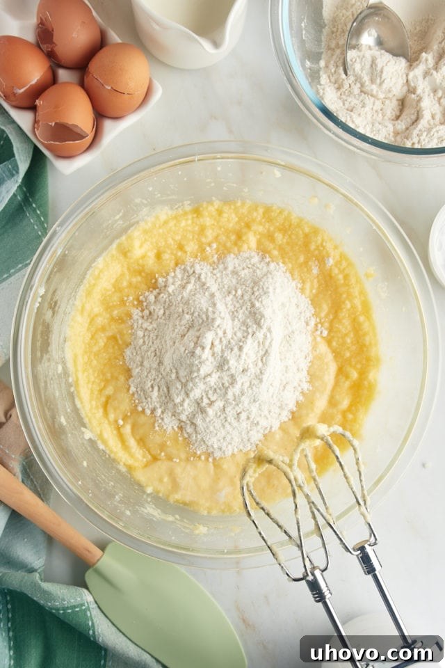 An overhead view of the flour mixture being added in portions to the wet cake batter in a glass mixing bowl, ensuring gradual and even incorporation.