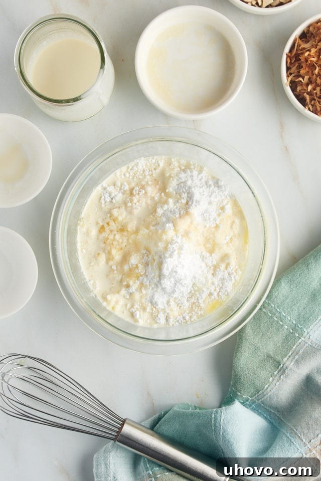 An overhead view of confectioners' sugar, melted butter, vanilla extract, almond extract, and heavy cream gathered in a glass mixing bowl, forming the base for the sweet glaze of the Louisiana Crunch Cake.