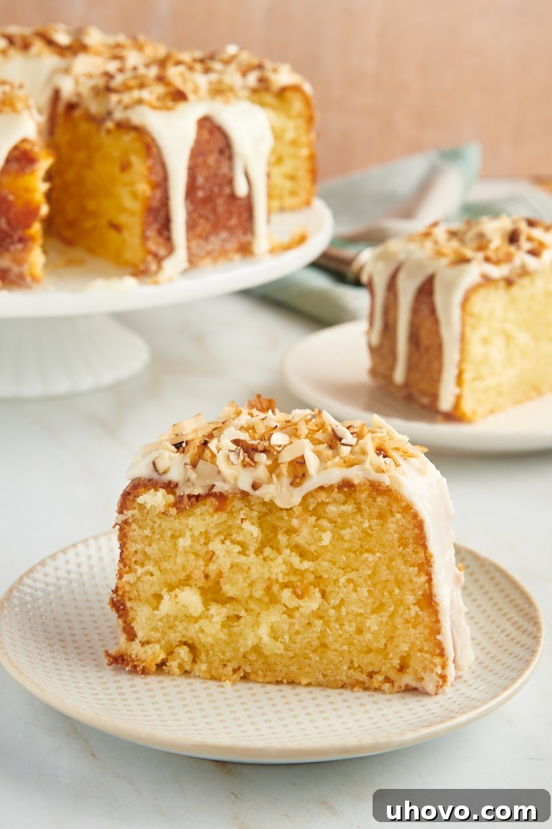 A perfectly golden slice of Louisiana Crunch Cake, showcasing its tender crumb, rich glaze, and abundant coconut and almond topping, resting on a charming white and beige plate with the rest of the cake in the soft-focused background.