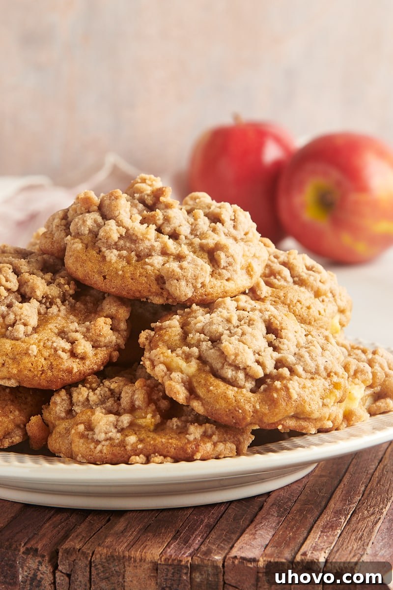 Crumb-topped apple cookies piled on a plate with two apples in the background.