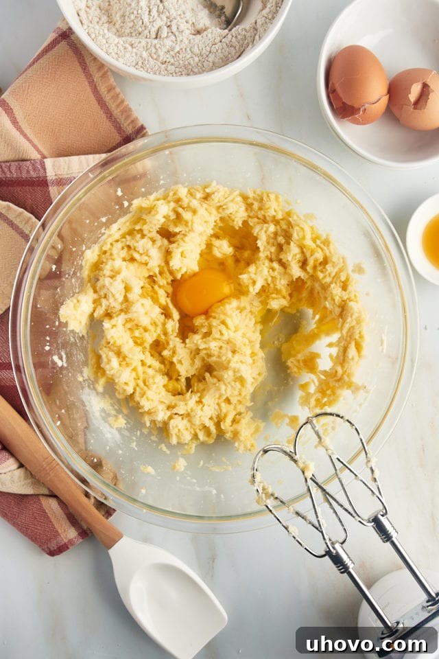 An overhead view captures an egg being carefully cracked into the creamy mixture of butter and sugars in a large mixing bowl, integrating the wet ingredients.