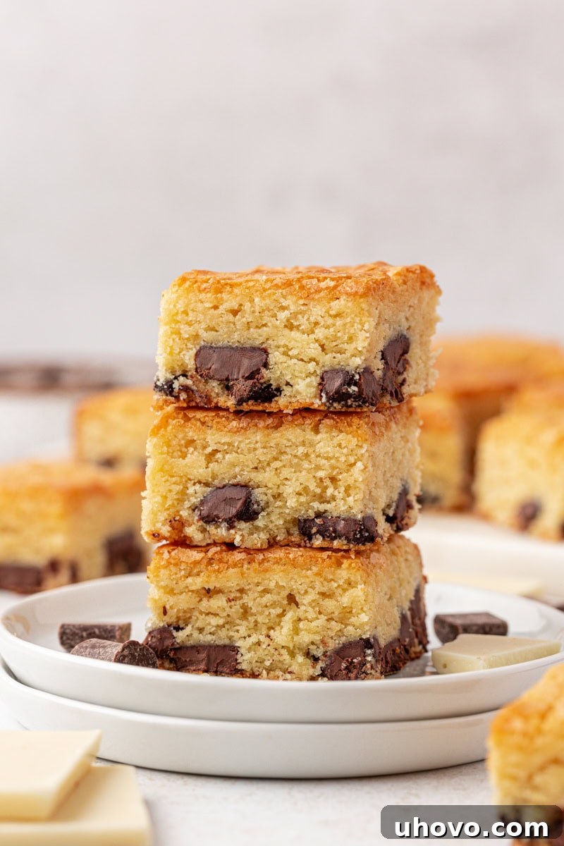 A stack of three decadent white chocolate brownies, featuring a beautiful golden crust and visible chocolate chunks, presented elegantly on a white plate with more brownies blurred in the background, suggesting abundance.