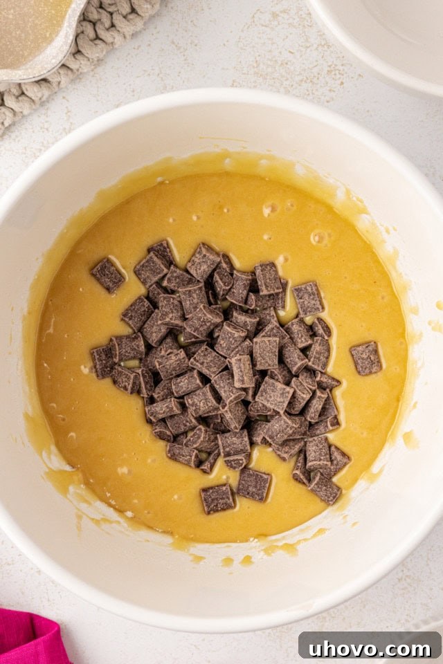 An overhead view of generous semisweet chocolate chunks being folded into the white chocolate brownie batter in a mixing bowl with a spatula.