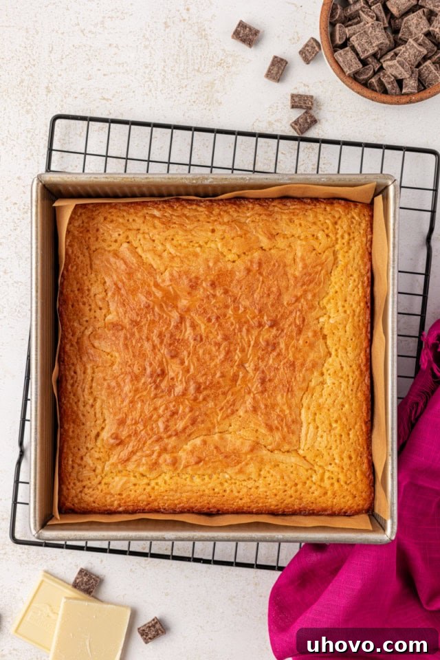 An overhead view of a freshly baked batch of golden-brown white chocolate brownies cooling in their square baking pan on a wire rack.