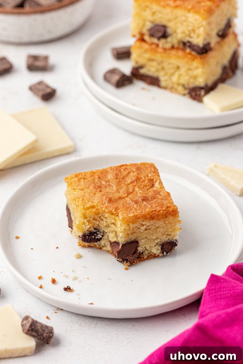 A single white chocolate brownie with a bite already taken out, perfectly showcasing its moist interior and chocolate chunks, served on a simple white plate with more brownies in the background.