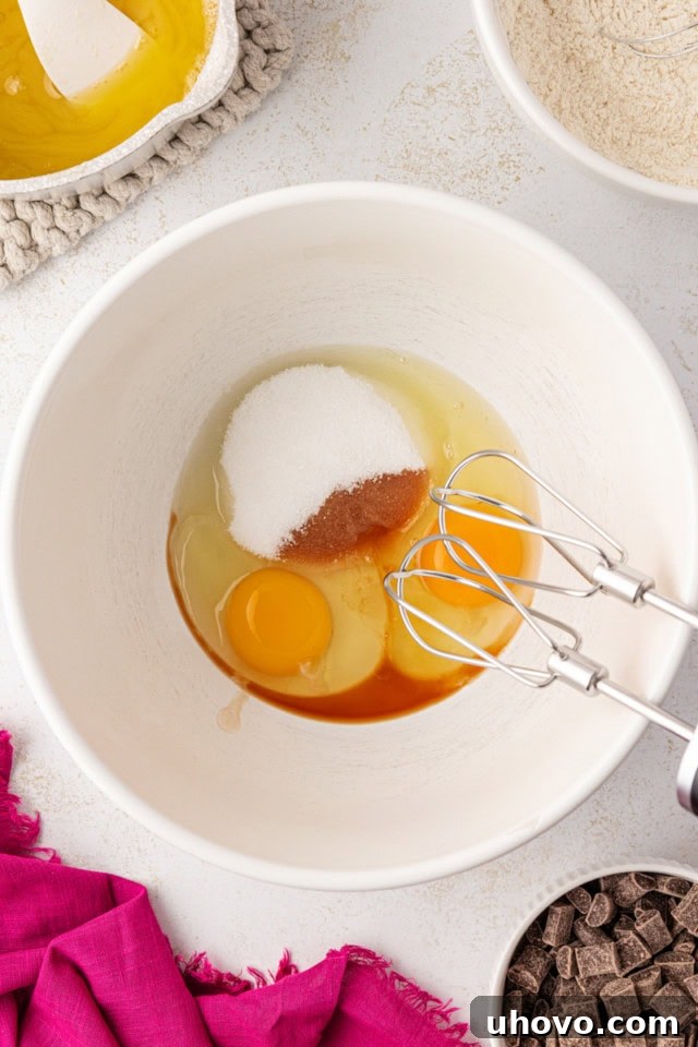 An overhead view of eggs, granulated sugar, and vanilla extract vigorously beaten together in a white mixing bowl to create a light and airy mixture.