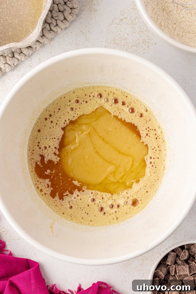 An overhead view as the slightly cooled melted white chocolate and butter mixture is carefully poured into the beaten egg and sugar mixture in a white mixing bowl.