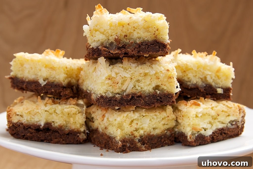 A stack of freshly baked Coconut Bars with Chocolate Shortbread Crust, each piece perfectly cut and displaying the golden coconut topping over the dark chocolate base, on a white serving pedestal.