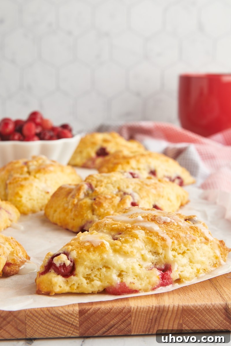cranberry orange scones on parchment paper on top of a wooden cutting board with a bowl of cranberries and a red mug in the background