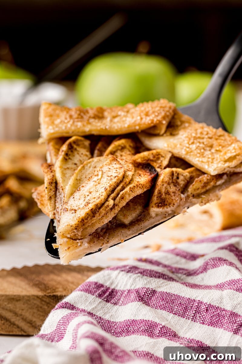 a slice of apple galette on a pie server with more pie and apples in the background