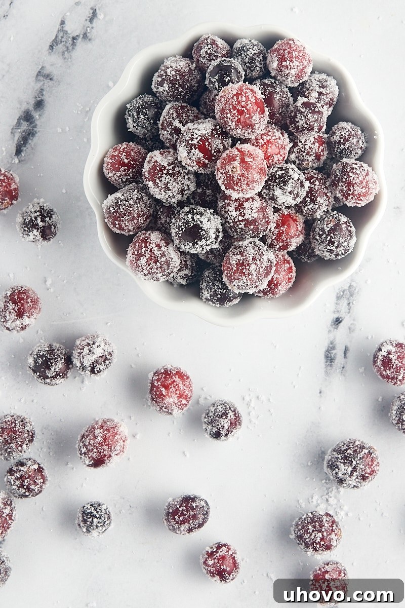 Overhead view of glistening sugared cranberries elegantly presented in a white bowl, with additional berries scattered around.