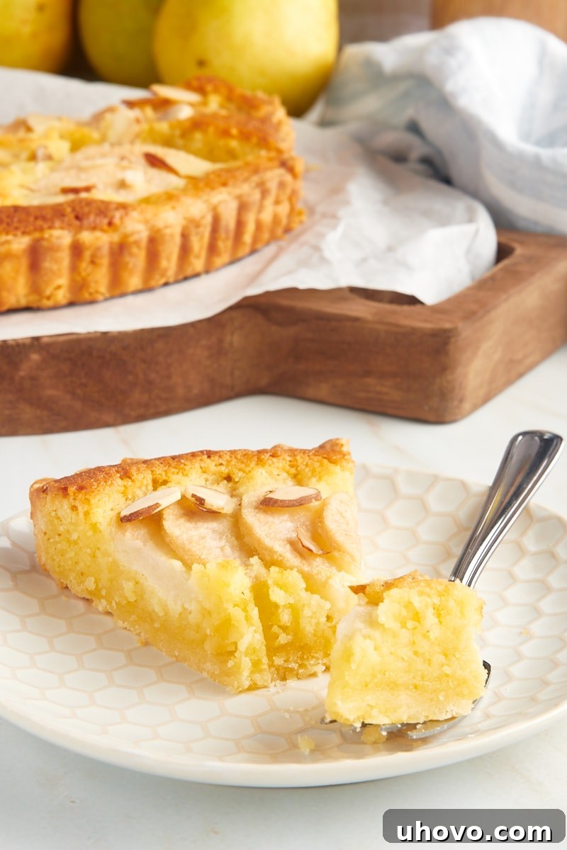 A close-up shot of a single slice of pear frangipane tart on a plate, with a fork gently lifting a piece, emphasizing the delicate layers and texture.