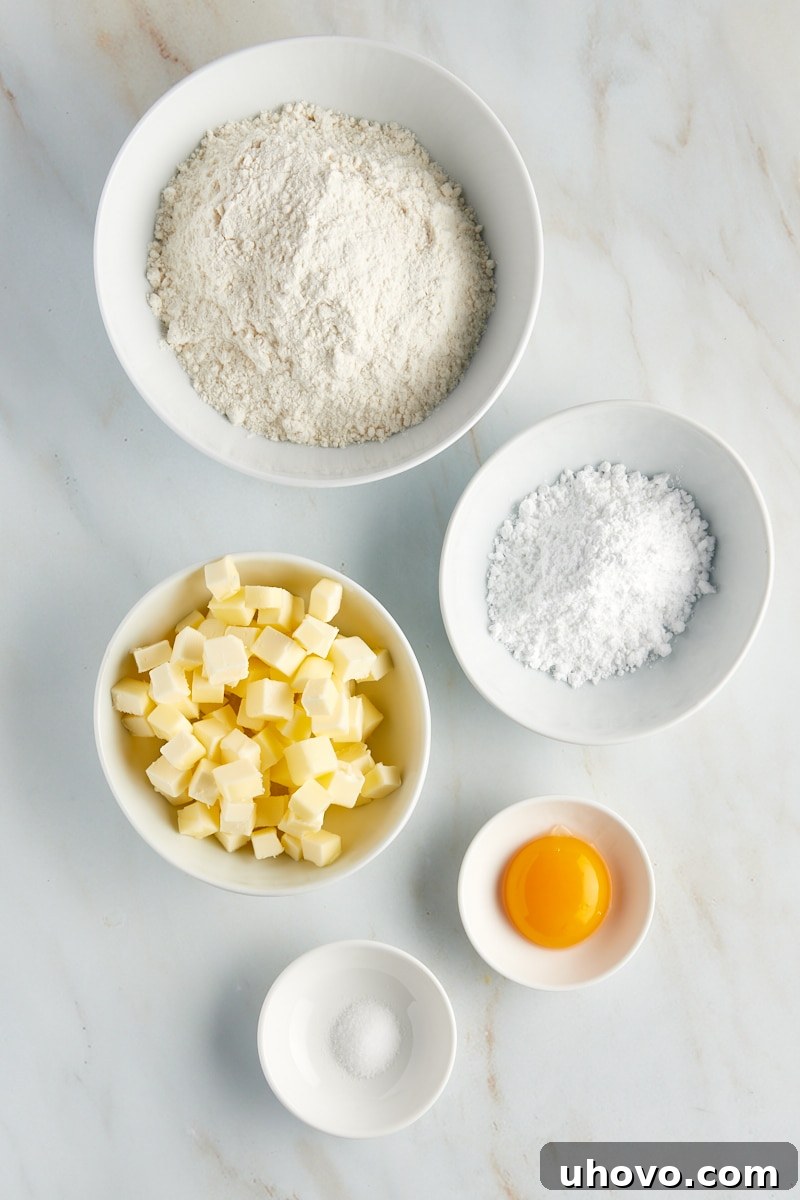 Overhead shot of all dry and wet ingredients for the tart crust, including flour, confectioners' sugar, salt, cubed butter, and an egg yolk in separate bowls.