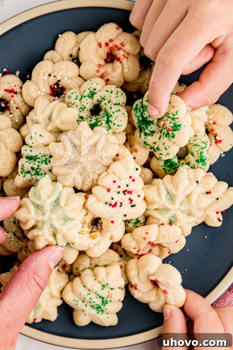 An overhead view of three hands reaching for intricately shaped spritz cookies from a plate brimming with them, highlighting their inviting appeal.