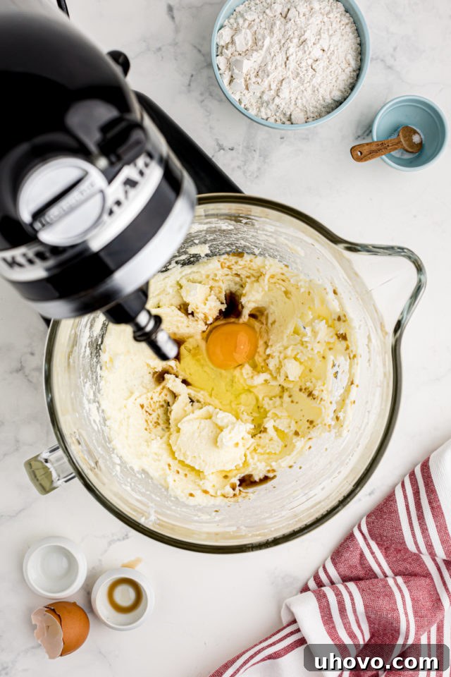 An overhead view of a large egg, vanilla extract, and almond extract just added to the creamed butter and sugar mixture in the stand mixer bowl, ready to be combined.