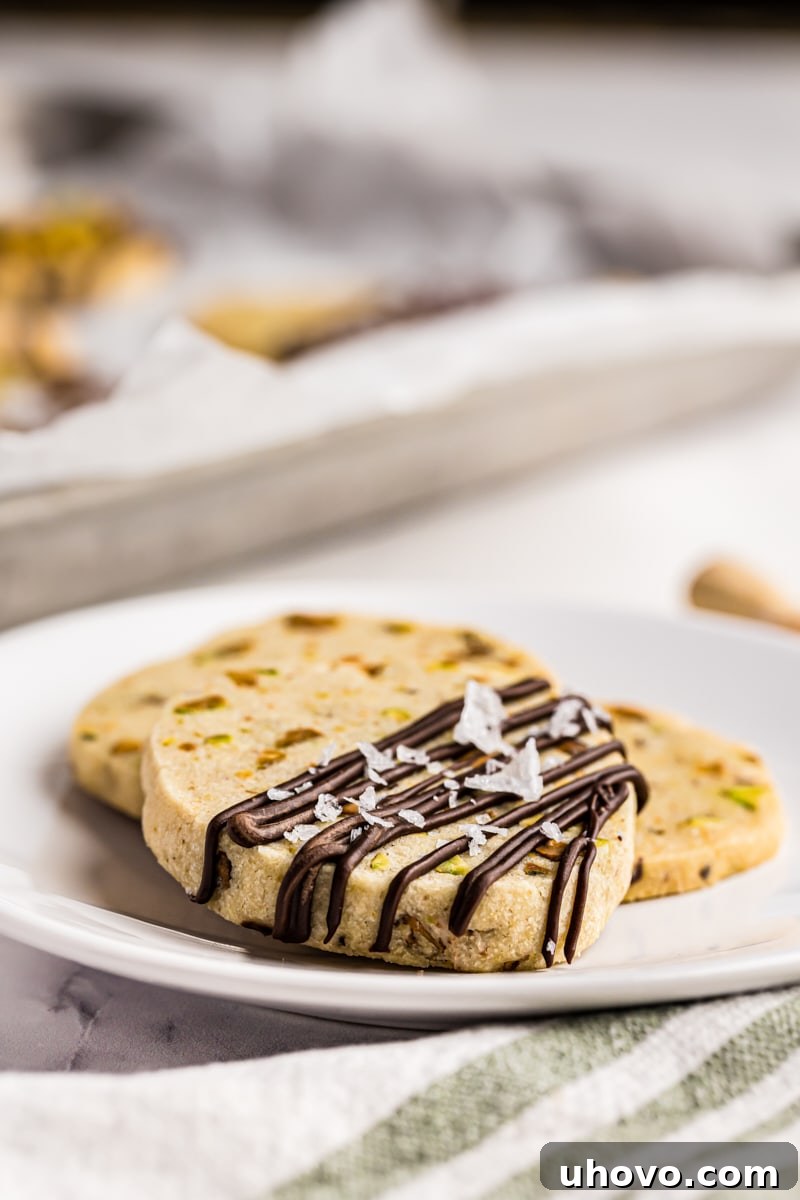 three pistachio cookies on a white plate with more cookies in the background