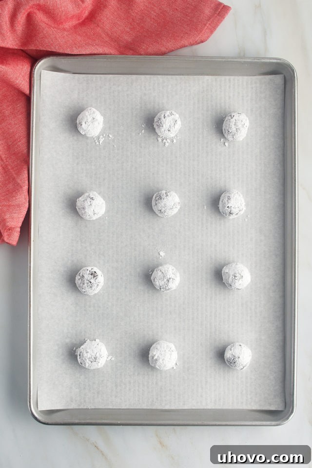 Chocolate crinkle cookie dough balls, thoroughly coated in powdered sugar, arranged on a parchment-lined baking sheet, ready for the oven.