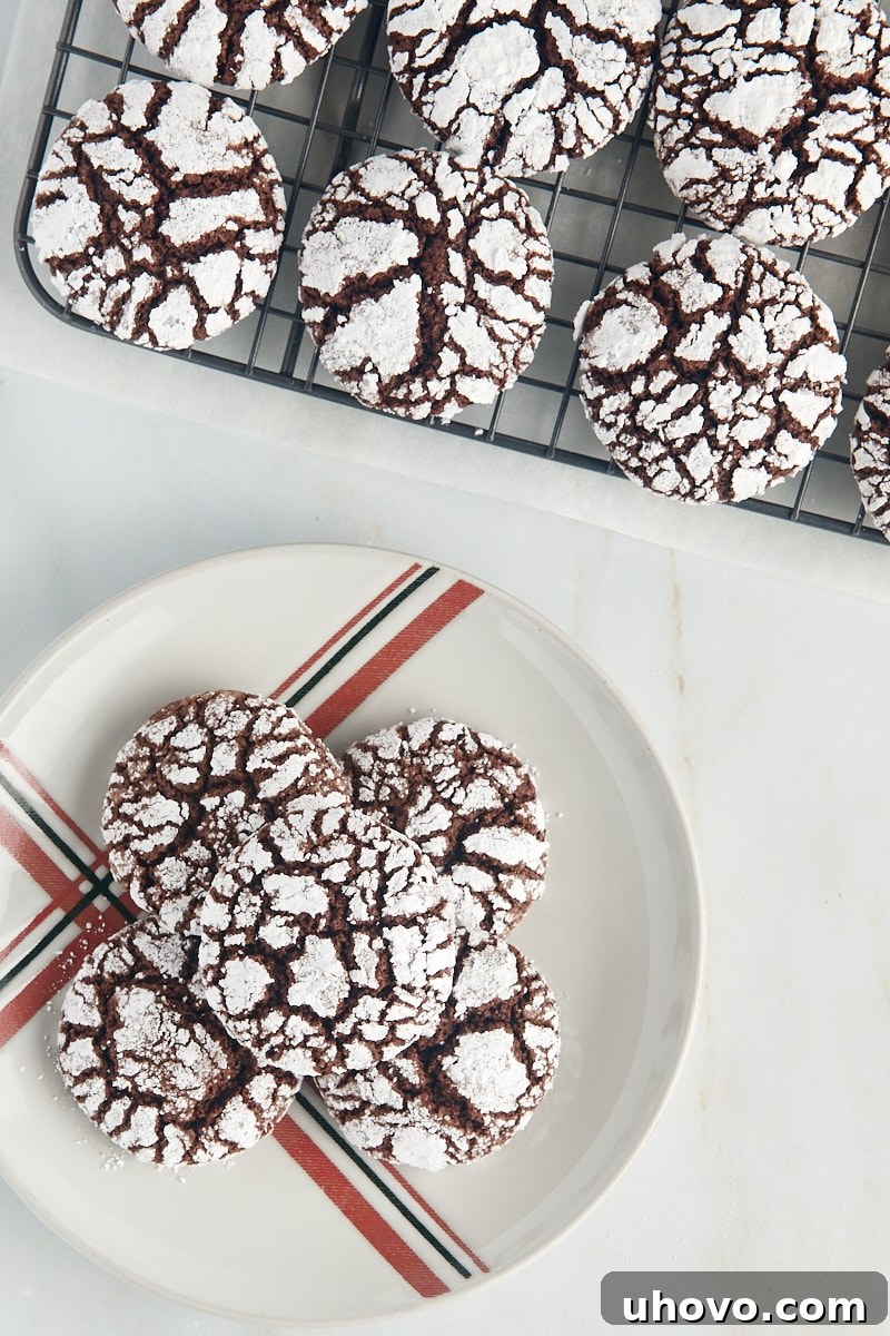 Overhead view of several chocolate crinkle cookies on a red and green striped plate, with more cookies cooling on a wire rack beside it, suggestive of holiday baking.