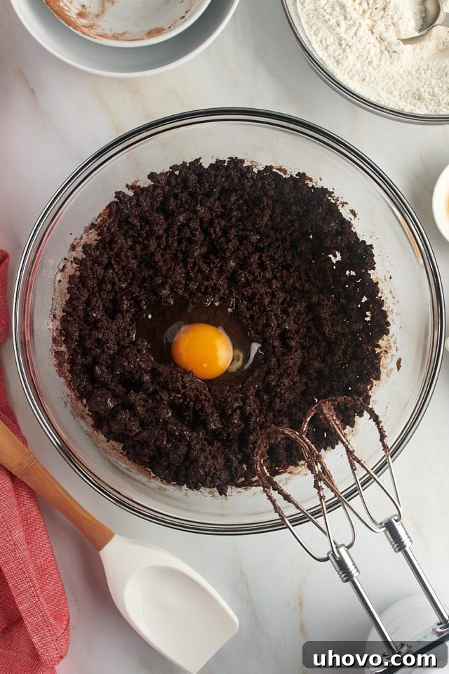 Overhead view of a single egg being added to the partially mixed chocolate crinkle cookie dough in a glass bowl, ready to be incorporated.