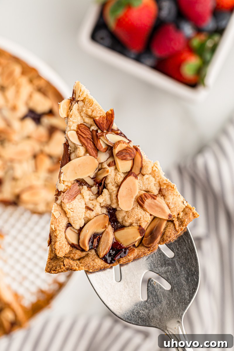 A slice of Italian Jam Crostata resting on a silver pie server, with the remainder of the tart in its pan and a bowl of fresh berries blurred in the background, suggesting a delightful serving.