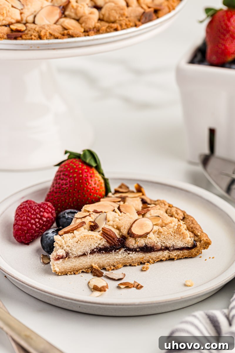 A delightful slice of Italian Jam Crostata on a pristine white plate, beautifully garnished with fresh berries (strawberries and blueberries), showcasing its homemade charm.