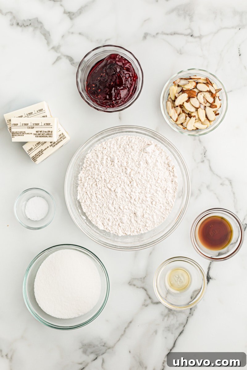 Overhead view of neatly arranged ingredients for Italian Jam Crostata on a light-colored surface, including butter, sugar, flour, extracts, jam, and sliced almonds.