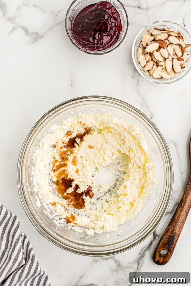 Overhead view of vanilla and almond extracts being added to creamed butter and sugar in a clear glass mixing bowl, illustrating the first step of combining wet ingredients.