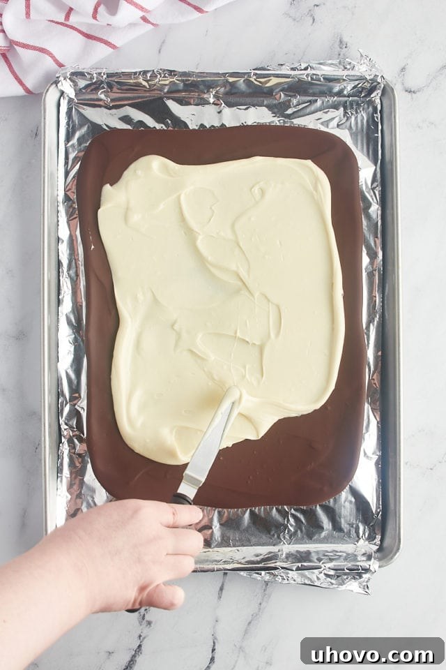 Overhead view of melted white chocolate being gently spread over the chilled dark chocolate layer on a foil-lined baking pan.